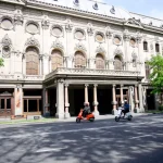 The white facade of the Rustaveli National Theatre on a sunny day, with two people riding mopeds in front of the building