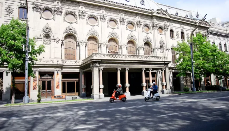 The white facade of the Rustaveli National Theatre on a sunny day, with two people riding mopeds in front of the building