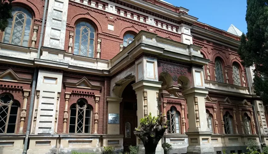 The ornate red brick facade of the State Silk Museum in Tbilisi, Georgia, featuring arched windows and an inviting entrance, bathed in sunlight.