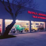 The exterior of the Tbilisi Auto Museum at dusk, showcasing a collection of vintage and classic cars through large illuminated display windows, with the museum's neon sign visible in Georgian and English.