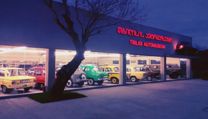 The exterior of the Tbilisi Auto Museum at dusk, showcasing a collection of vintage and classic cars through large illuminated display windows, with the museum's neon sign visible in Georgian and English.