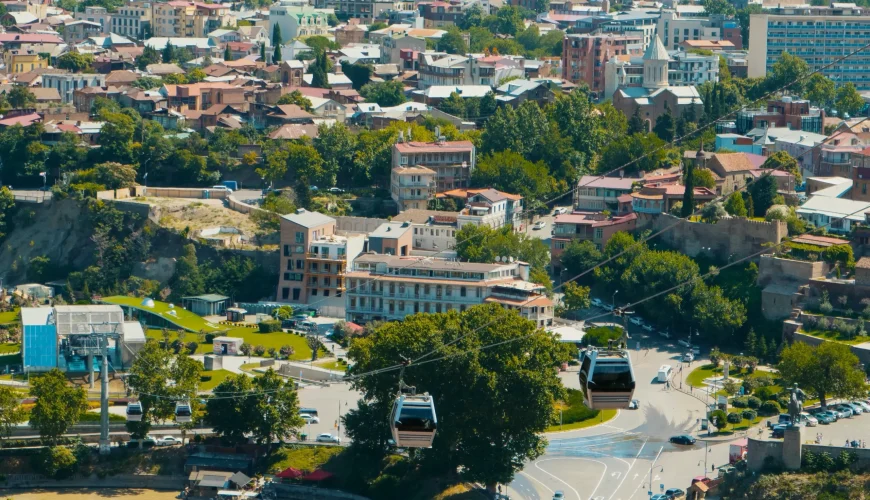 An aerial view of Tbilisi with the Kura River, a modern bridge, and cable cars moving through the city skyline
