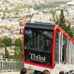 A red and black funicular car on a steep track with the city of Tbilisi and forested hills in the background