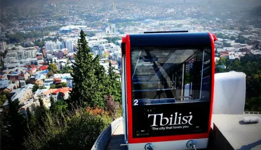 A red and black funicular car on a steep track, with a panoramic view of the Tbilisi city skyline and mountains in the background