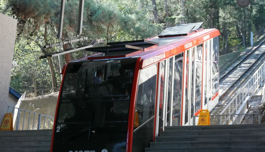 The Tbilisi Funicular car arriving at a station, with a staircase leading down and green trees visible in the background