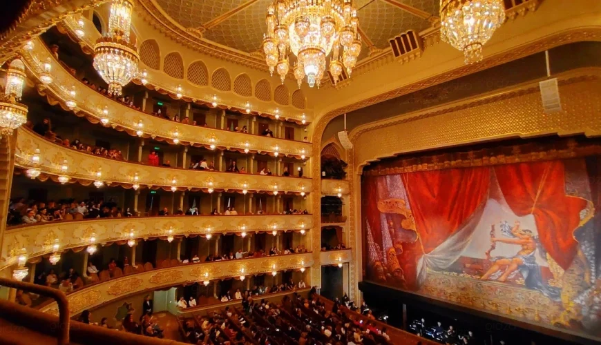 An ornate interior shot of the Tbilisi Opera and Ballet Theatre with gold balconies, a large chandelier, and a stage with a red curtain and mural