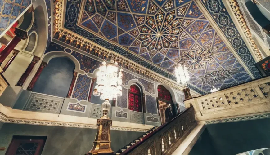 The ornate interior staircase of the Tbilisi Opera and Ballet Theatre with a mosaic ceiling and a large, hanging chandelier